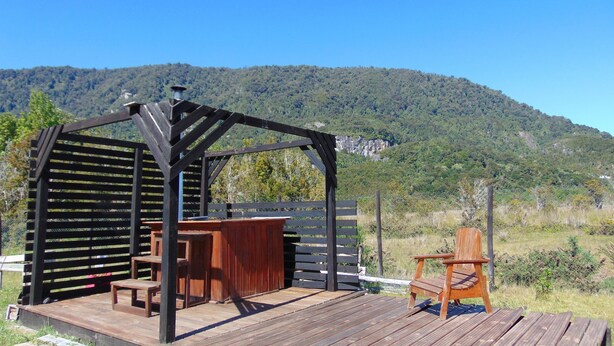 Een houten terras met een pergola, buitenmeubels en uitzicht op een bebost gebergte.