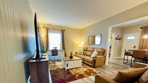 Ein Wohnzimmer mit einer Couch, einem Couchtisch und einem Fernsehschrank. Ein Fenster mit Vorhängen und ein kleiner Weihnachtsbaum sind ebenfalls zu sehen.
