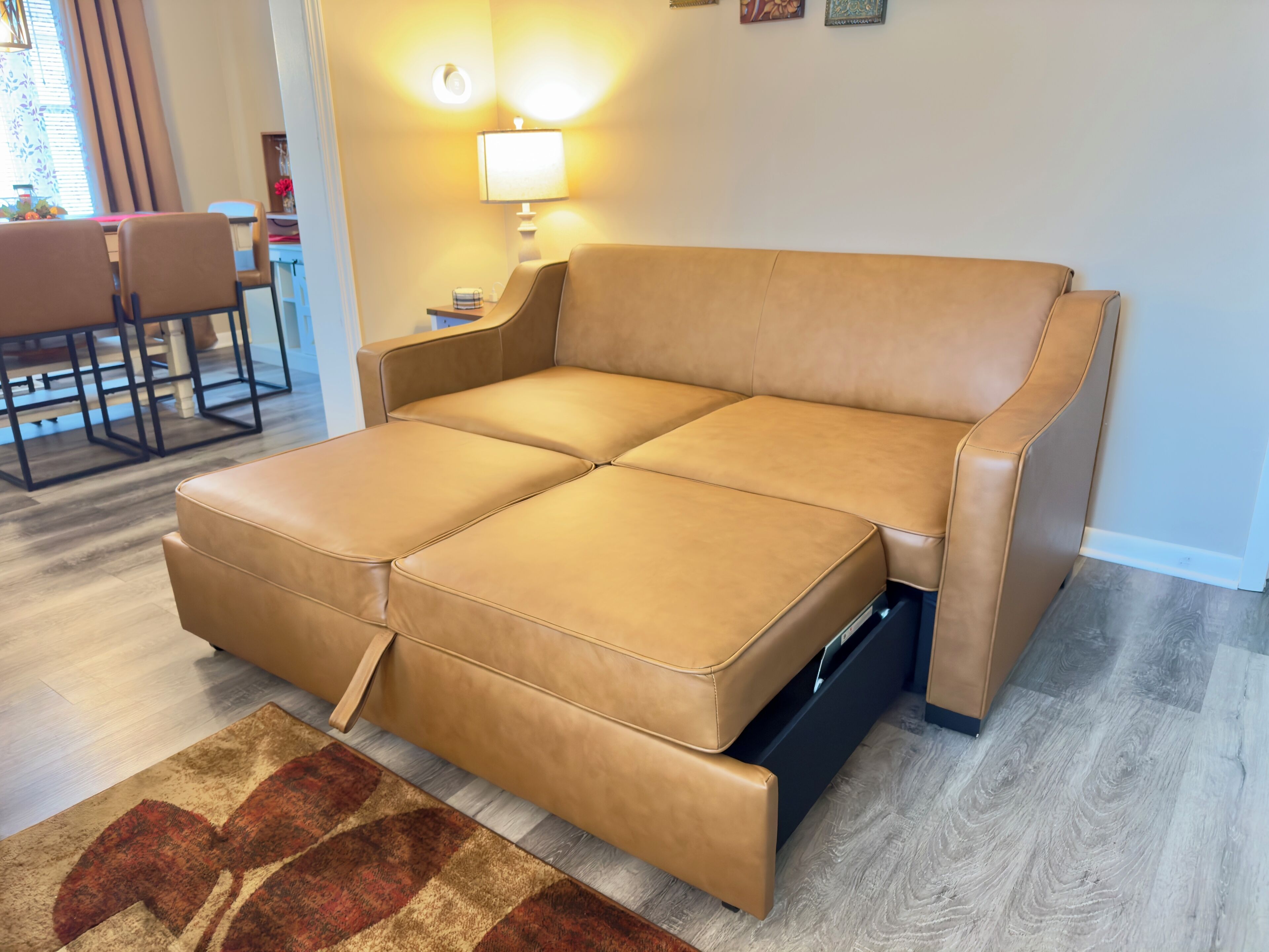 A tan leather sectional sofa with a pull-out bed in a living room with wooden flooring and a patterned rug.