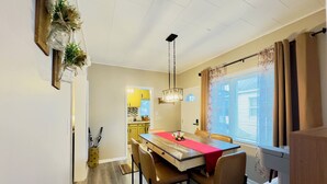 Ein Essbereich mit einem Holztisch, Stühlen und einer roten Tischdecke. Rechts befindet sich ein Fenster mit Vorhängen und eine Kaffeemaschine.