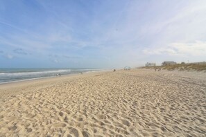 Ein weiter, sandiger Strand mit sanften Wellen, einigen Personen und Gebäuden im Hintergrund.
