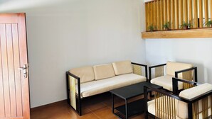 Une pièce comprenant un canapé, deux fauteuils, une table basse, un ventilateur de plafond et une étagère en bois à lattes sur laquelle sont disposées des plantes.