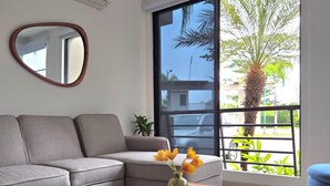 Ein modernes Wohnzimmer mit grauem Sofa, einem Holztisch mit Blumenvase und einem großen Fenster mit Blick auf Palmen und einen Balkon.