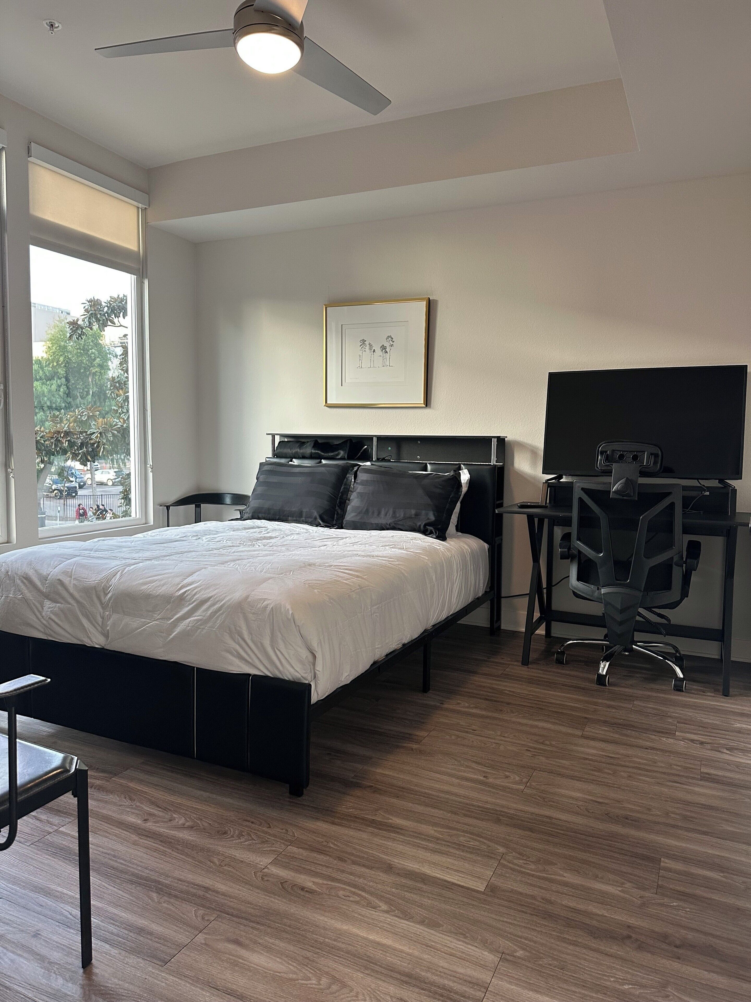 Ein modernes Schlafzimmer mit Bett, Schreibtisch, Stuhl, Deckenventilator und einem Fenster mit Blick auf die Straße.