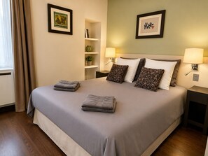 A neatly made bed with pillows and folded towels, a bedside table with a lamp, a shelf with books and a plant, and two framed pictures on the wall. - L'appartement des Archives (Paris)