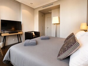 Quarto de hotel com uma cama, televisão sobre um suporte, uma cadeira, escrivaninha e abajur.