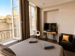 Habitación de hotel con cama, televisor en un mueble, una silla, ventana con cortinas y vistas a edificios.