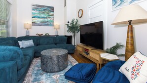 A living room with a teal sofa, a round ottoman, a TV, and a lamp.