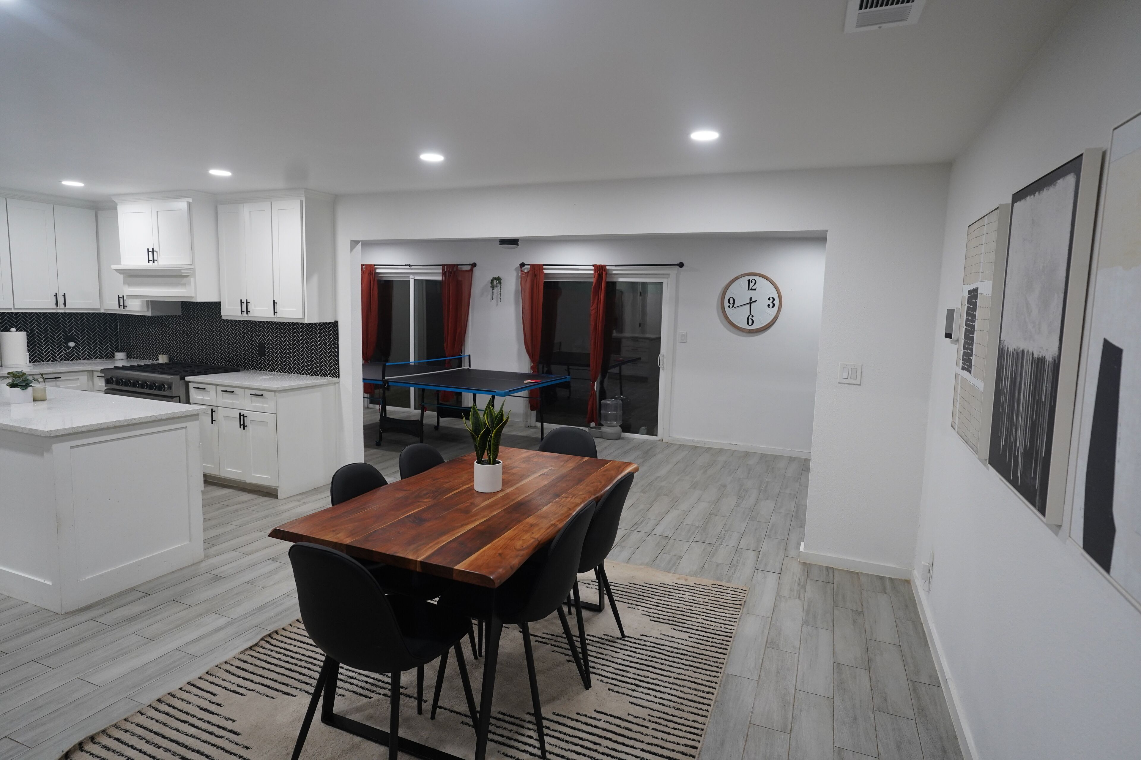 A modern kitchen with white cabinets, a wooden dining table, and a ping pong table in the background.