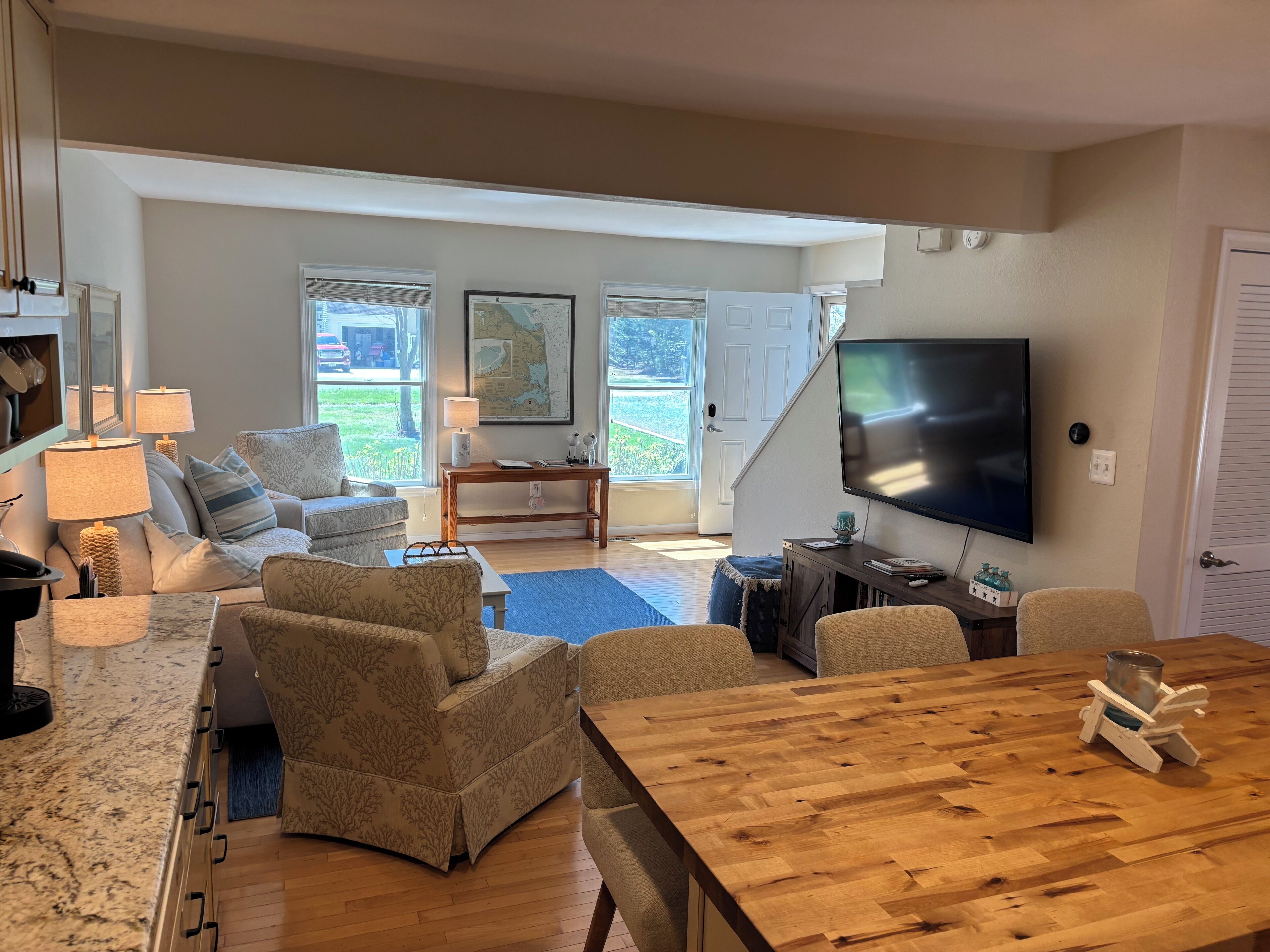 A spacious living room with a large wooden table, comfortable seating, a flat-screen TV, and a view of the outdoors through large windows.