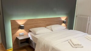 A neatly made bed with white linens and a folded towel, a bedside lamp, and a wall-mounted air conditioner.