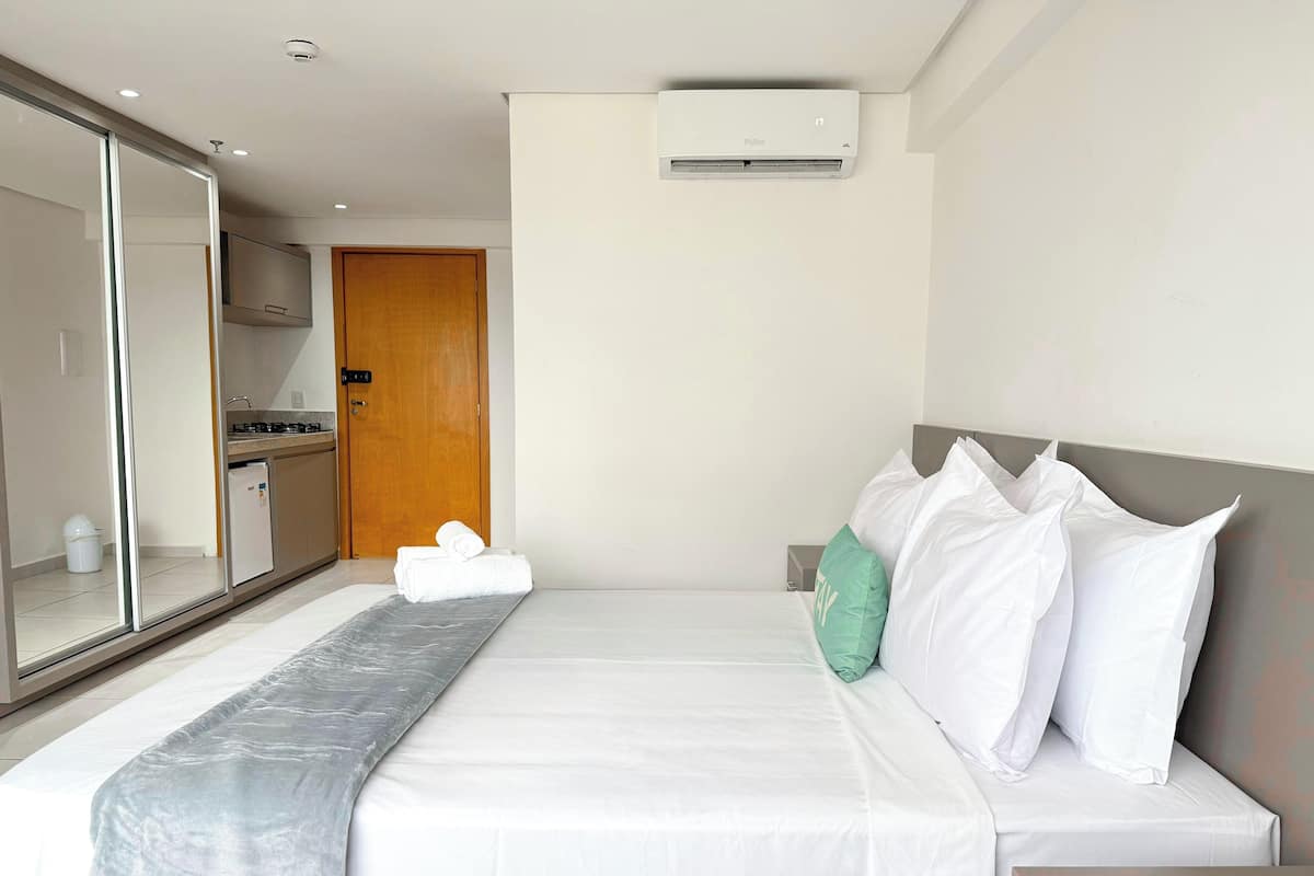 A modern hotel room with a bed, a nightstand, a mirror, a wardrobe, a kitchenette, and an air conditioning unit.