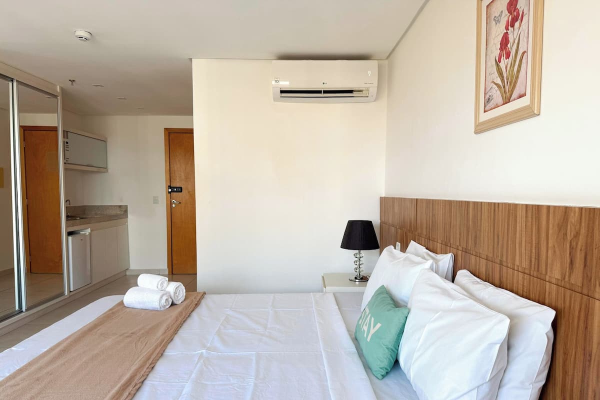 A hotel room with a bed, a bedside table with a lamp, a wall-mounted air conditioner, and a framed botanical illustration.