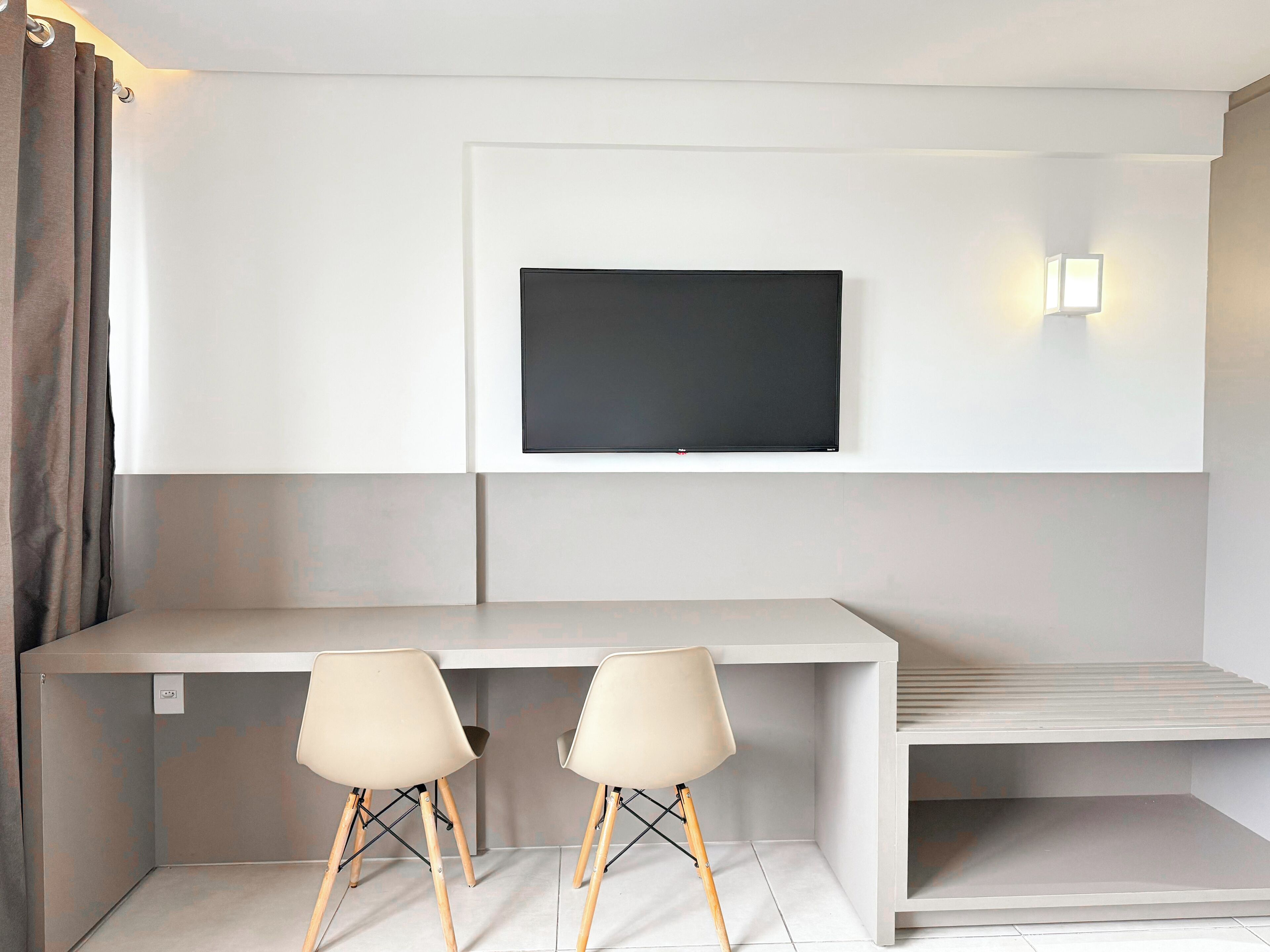 Une chambre moderne avec un téléviseur à écran plat fixé à un mur blanc, un bureau blanc et deux chaises aux pieds en bois.