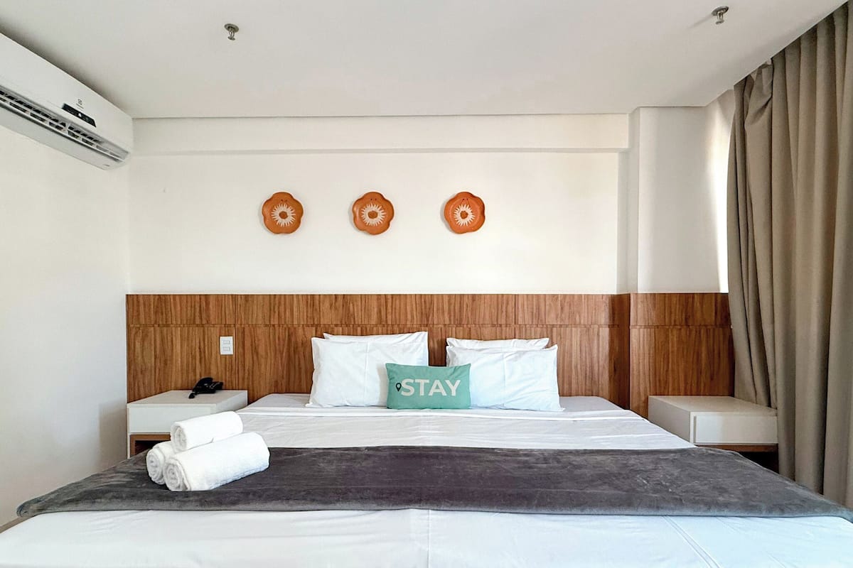 A hotel room with a wooden headboard, a bed with white bedding, a 'STAY' pillow, and a wall-mounted air conditioner.