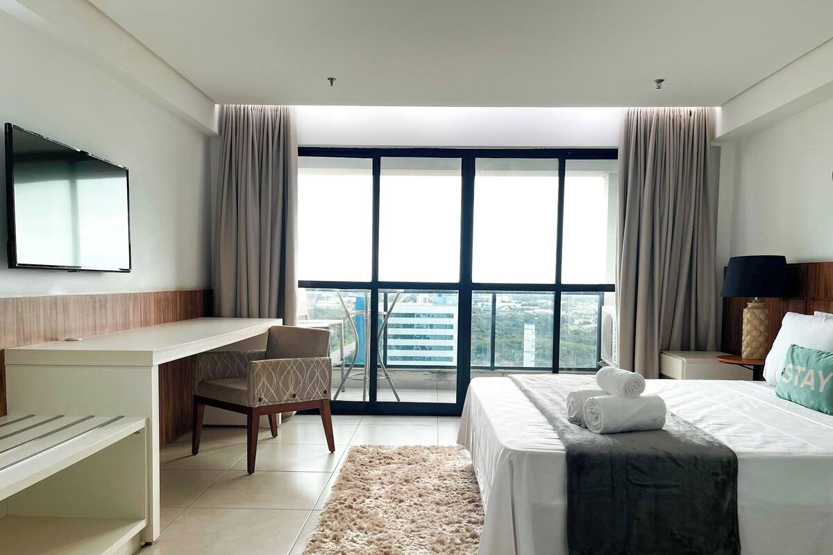 A hotel room with a bed, a desk, a chair, a TV, and a large window with curtains.