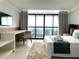 Una habitación de hotel con cama, escritorio, silla, televisor y un ventanal con cortinas.