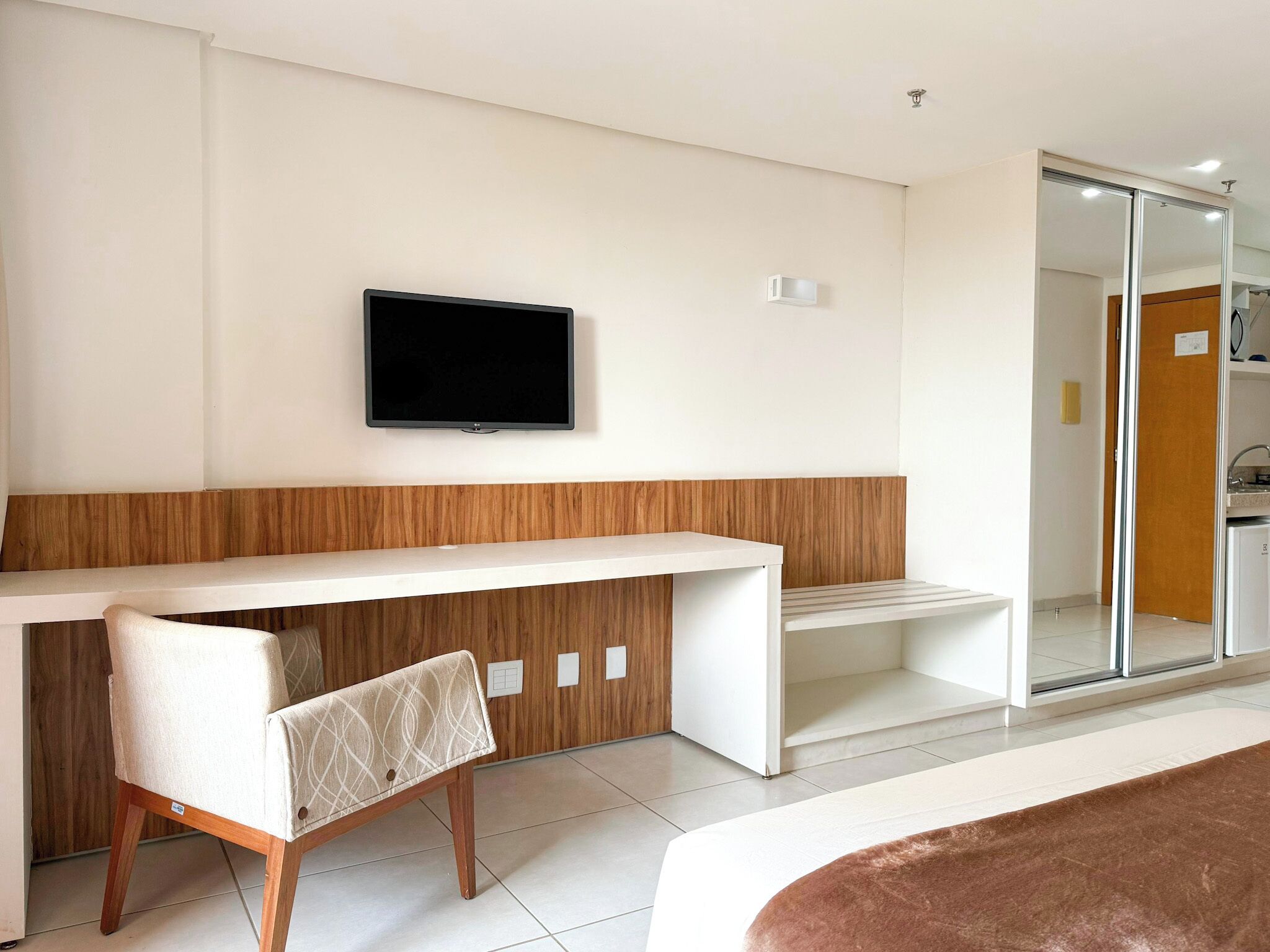 Une chambre d’hôtel moderne avec un bureau blanc, un téléviseur à écran plat fixé au mur, un mur d’accent en bois, une chaise avec un coussin à motifs et un grand miroir.