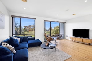 A modern living room with a sectional sofa, a coffee table, a TV, and large windows offering a view of the outdoors. - Magic Driftwood (Torquay)
