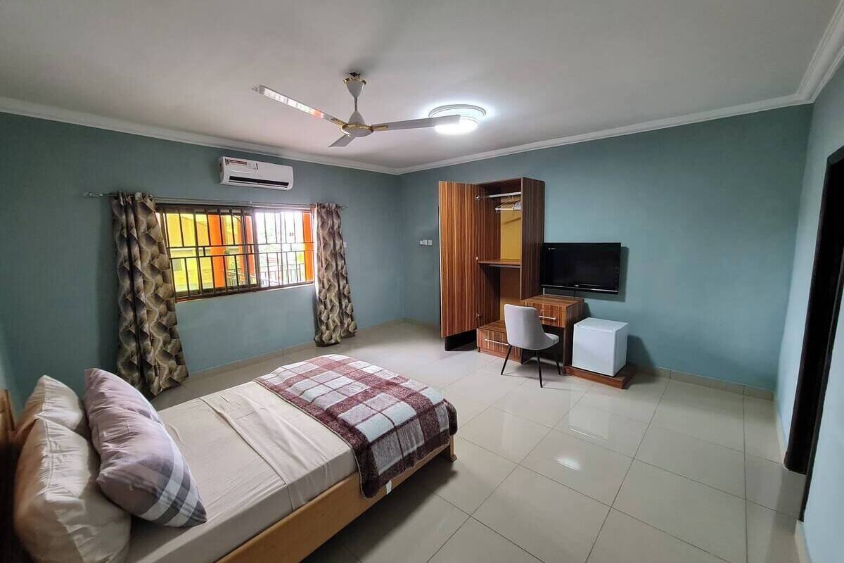 A bedroom with a bed, a desk, a chair, a television, and an air conditioning unit.