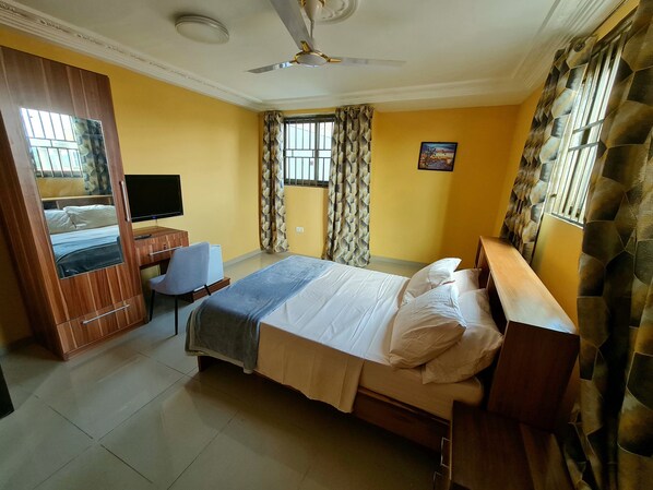 A bedroom with a bed, a desk, a chair, a television, and a ceiling fan. - The Charways Lodge (Accra)