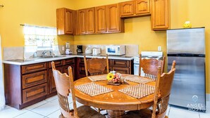 A kitchen with wooden cabinets, a stainless steel refrigerator, a microwave, and a stove. There is a dining table with chairs and a vase of flowers. - Chalk Sound Beach Residences (Providenciales)