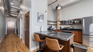 A modern kitchen with stainless steel appliances, a wooden floor, and a bar area with two stools. - Cooperage 214 | Mid-century 1BD Loft | Peoria (Peoria)