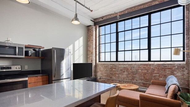 A modern kitchen with a large island, a refrigerator, and a microwave. There is a large window with a view of the sky. - Cooperage 214 | Mid-century 1BD Loft | Peoria (Peoria)