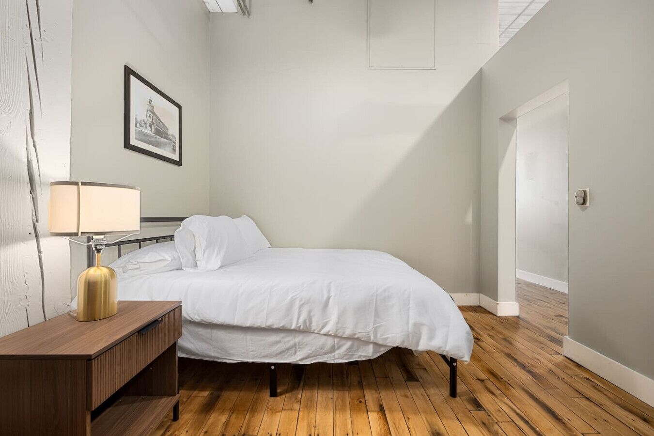 Un dormitorio con cama, mesita de noche, lámpara y un cuadro en la pared.