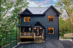 Una casa de dos pisos con fachada oscura, un porche con banca de madera y vistas al bosque circundante.