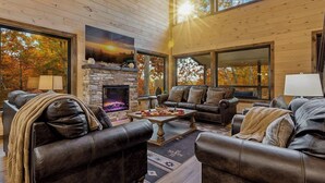 A cozy living room with leather sofas, a stone fireplace, and large windows offering views of autumn trees. - Gatlinburg Lux Cabin | Indoor Pool | Huge Arcade! (Gatlinburg)