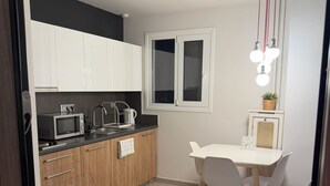 Een kleine keuken met witte kasten, een magnetron, een waterkoker en een eethoek met een witte tafel en stoelen.