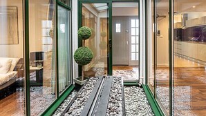 Un intérieur moderne avec une entrée dotée d’une porte en verre, un petit jardin intérieur agrémenté de topiaires sphériques, et une cuisine visible à travers le verre.