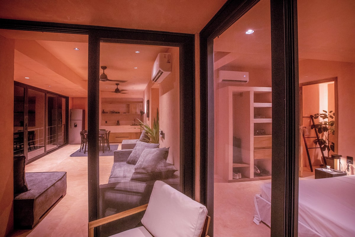 A modern hotel room with a glass partition, a sofa, a bed, and a dining area with a table and chairs.