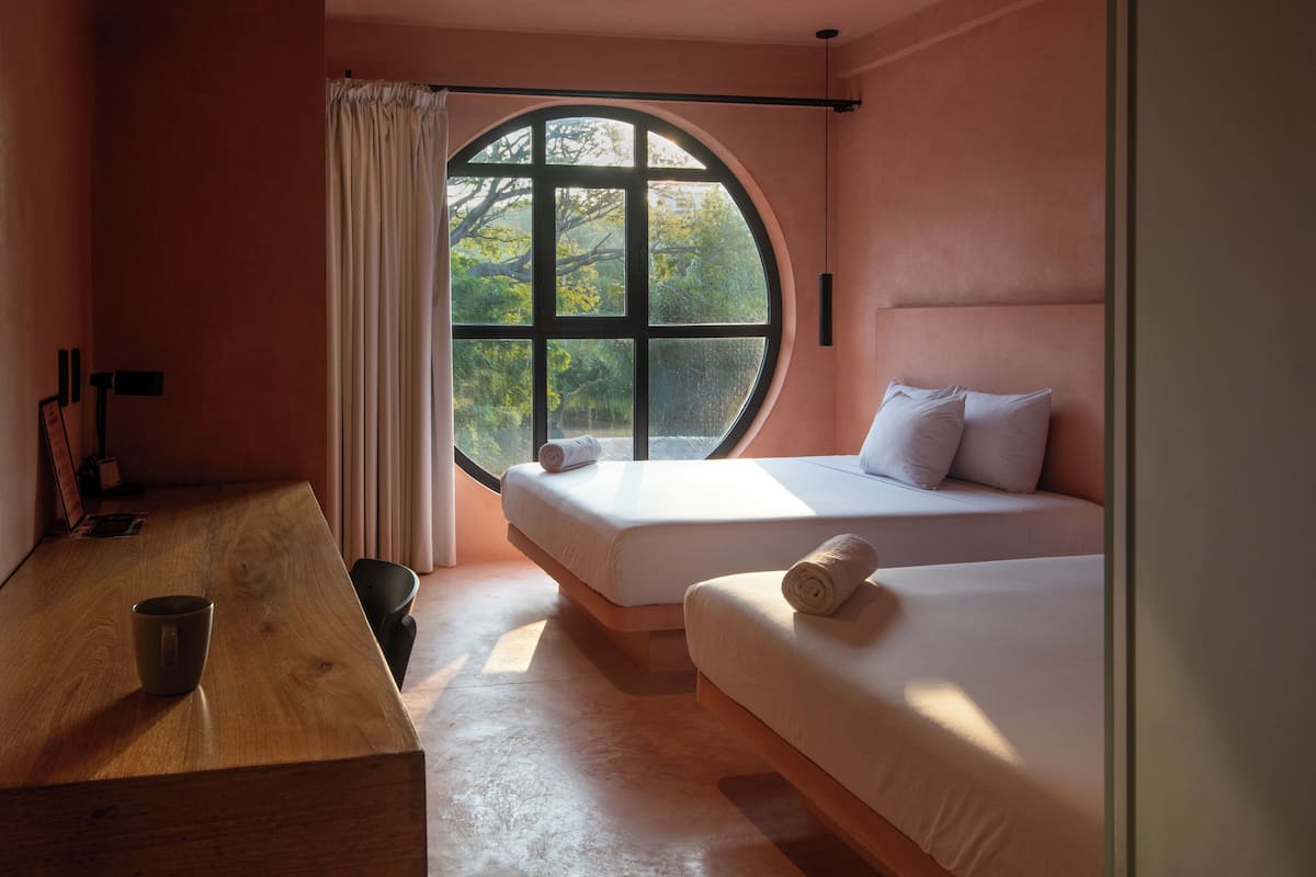 A room with a large circular window, two beds, a wooden table, and a chair.