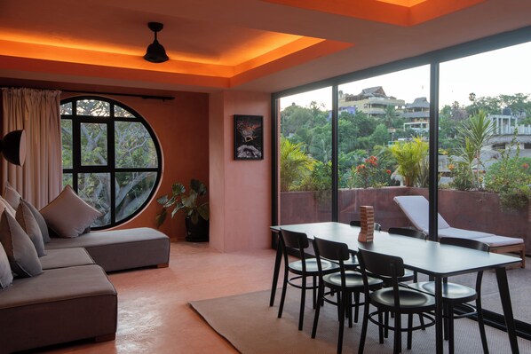 A modern living room with a large window, a sofa, a dining table with chairs, and a ceiling fan.