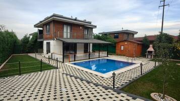 A modern two-story house with a swimming pool, surrounded by a patterned patio and a small garden.