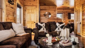 A cozy log cabin interior with a brown leather sofa, a round coffee table with snacks and drinks, and a wooden ceiling.