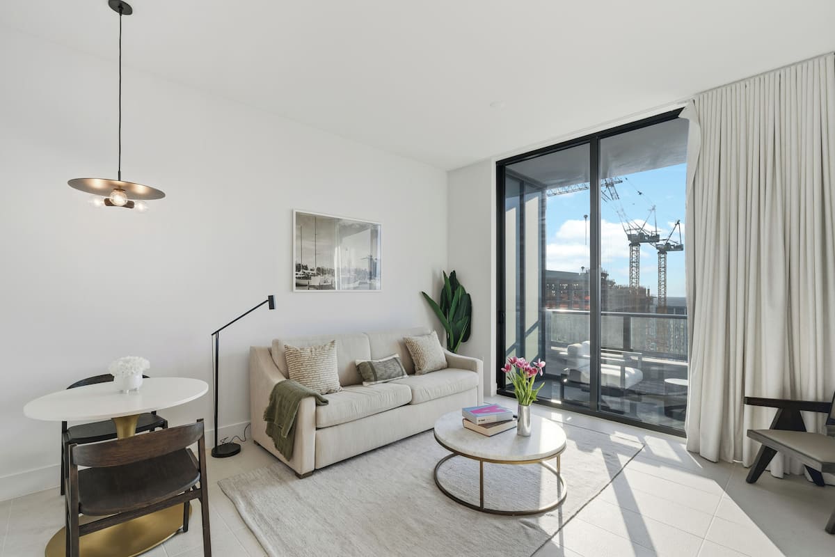 A modern living room with a sofa, a round coffee table, a floor lamp, and a large window with a view of the city.