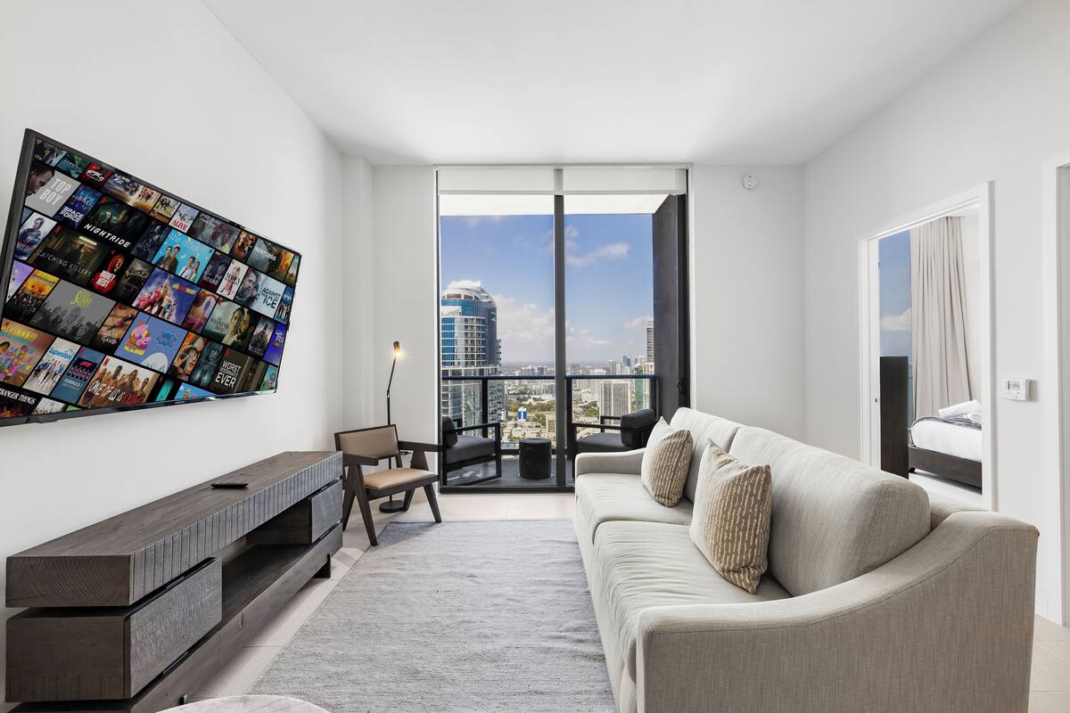 A modern living room with a large TV displaying movie posters, a sofa, a coffee table, and a view of the city through large windows.