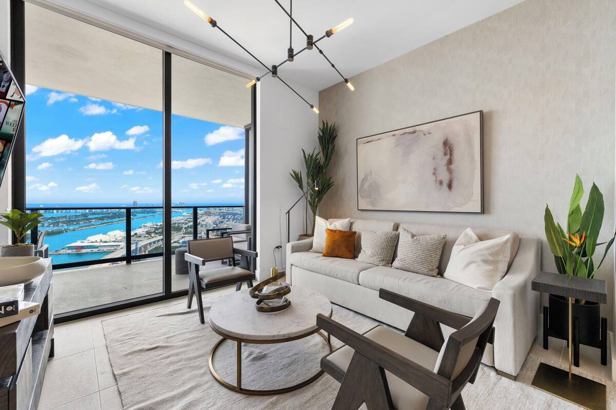 A modern living room with a sofa, coffee table, and chairs, a large window with a view of the ocean, and a painting on the wall.