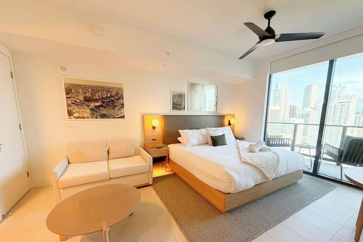 A modern hotel room with a large bed, a sofa, a small table, and a balcony with a city view.