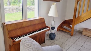 Huoneessa on piano, ja suuresta ikkunasta avautuu näkymä puutarhaan. Lisäksi huoneessa on lamppu ja puinen portaikko.