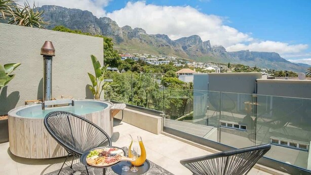 A rooftop terrace with a hot tub, two chairs, a table with food and drinks, and a view of mountains and a town.