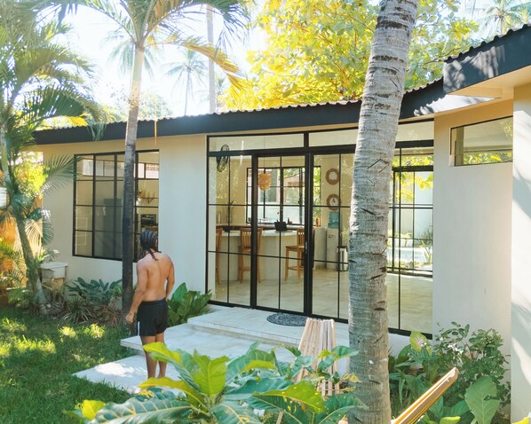 Una casa moderna con puerta de entrada de vidrio, un comedor con mesa y sillas, y un jardín con plantas tropicales.