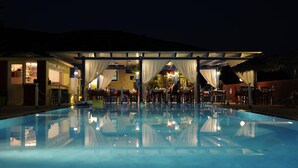 Vista noturna de uma área de piscina de hotel com um espaço de refeições coberto e um bar.