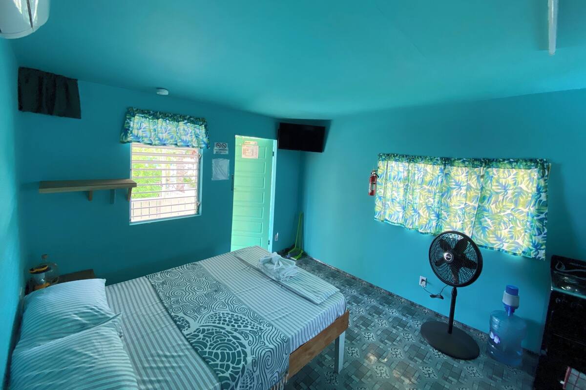 Une chambre avec un lit, une fenêtre avec des rideaux, un ventilateur et un distributeur d’eau.