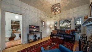 Ein Wohnzimmer mit einer schwarzen Ledersofa, einem blauen Sessel, einem Couchtisch aus Holz und einem großen Teppich. Ein Fernseher steht auf einem Schrank, eine Lampe ist vorhanden und das Fenster bietet Blick auf Bäume.