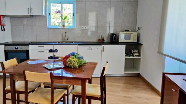 Cozinha compacta com armários brancos, uma mesa de jantar com quatro cadeiras e uma janela com vista para o verde.