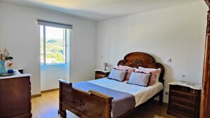 Quarto com cama de madeira, mesa de cabeceira, cômoda e janela com vista para o verde.
